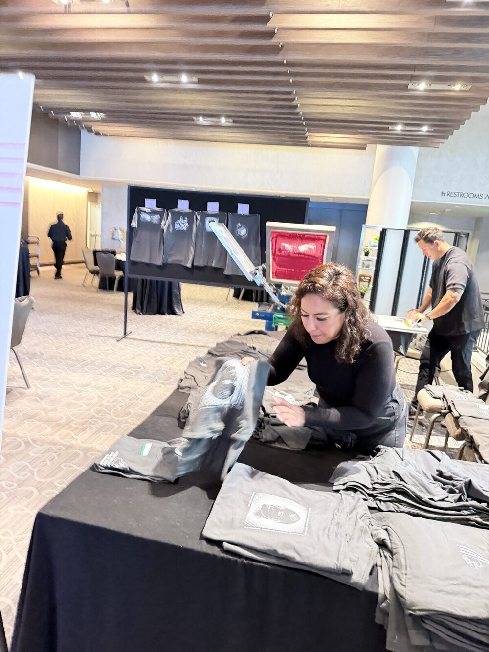 A crew member inspecting a freshly printed black shirt beside stacks of folded prints at an event table